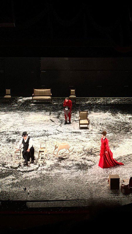 Вид сверху на сцену: несколько артистов в исторических костюмах, расставленные стулья и толстый слой бумажного снега — динамика сценического действия.