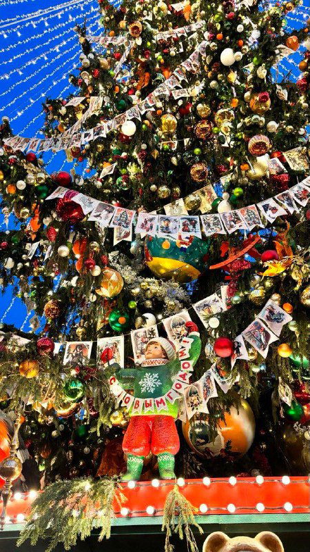 Крупный план ветвей и ретро-игрушек на елке: флажки, ватные персонажи и куклы в народных костюмах на фоне огней городской иллюминации.