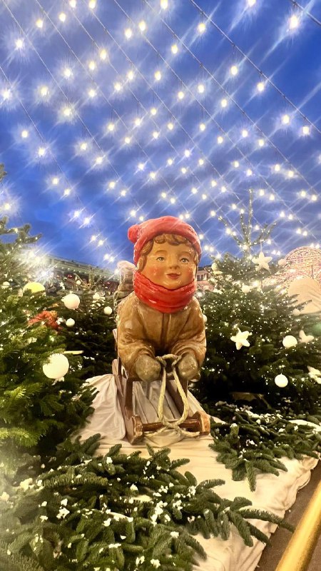 Фигурка в образе ребенка на праздничной площади под сеткой из гирлянд; декор создает иллюзию зимнего сказочного неба и фотозон.