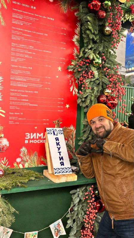 Пара на фоне стенда с программой проекта и рождественскими гирляндами; стенд указывает на маршрут и зоны фестиваля зимних локаций.