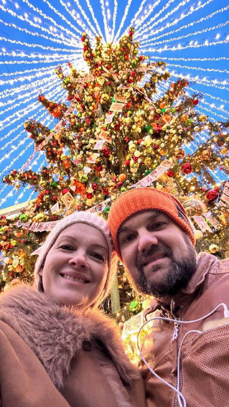 Селфи пары под высокой елкой, увитой гирляндами; видно слияние городской архитектуры и праздничной иллюминации в вечернем свете.