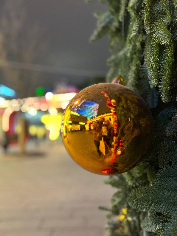 Крупный план золотого елочного шара, в отражении которого видны огни, люди и праздничные декорации городской улицы в вечернем освещении.