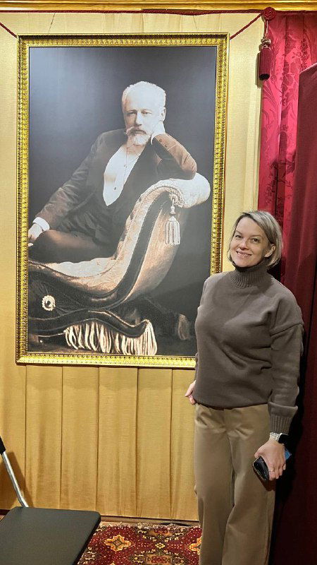 Посетительница рядом с большим портретом Петра Ильича Чайковского в экспозиции музея; интерьер, посетителька улыбается и держит телефон в руке.