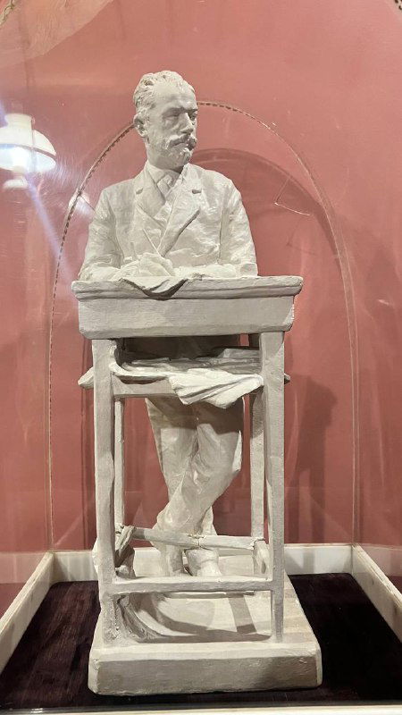 Белая гипсовая скульптура Чайковского в витрине, изображён сидящий композитор за столом; музейный экспонат, крупный план скульптурной фигуры.