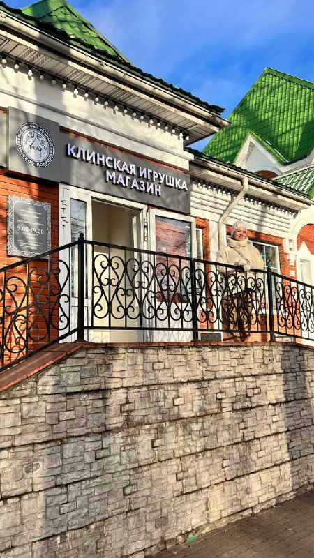 Фасад и вход в магазин «Клинская игрушка», кирпичная архитектура и кованое ограждение у ступеней; яркий кадр с уличной витриной на солнечном дне.