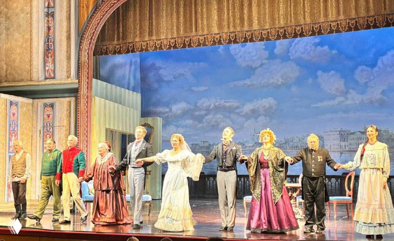 Актёры на поклоне на фоне сценографии: ансамбль в исторических костюмах завершает представление, публика аплодирует в зале.