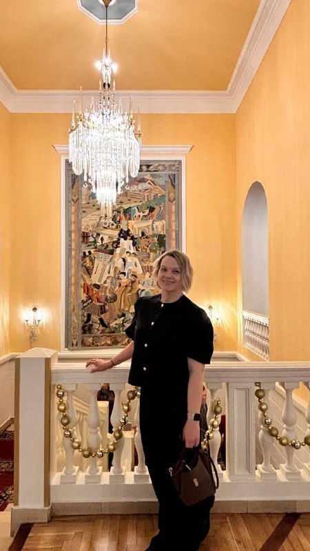 Посетительница театра позирует на лестнице фойе у декоративной стены и люстры: интерьер Малого театра с элементами оформления и праздничной гирляндой.