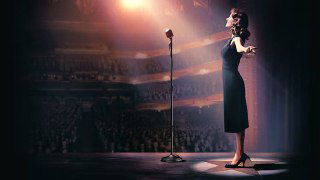 Героиня сериала на сцене большого театра или клуба, одиночный микрофон, яркий свет прожекторов и пустые ряды в полумраке зала