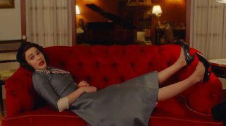 Героиня лежит на красном диване в ретро-костюме, кадр показывает сочетание юмора и камерной бытовой сцены с акцентом на одежду