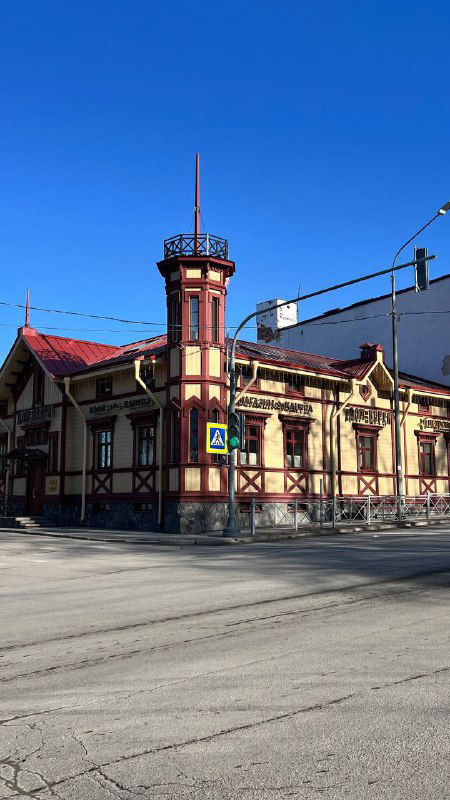 Угловой исторический дом с декоративным фасадом на пересечении улиц; пустой перекрёсток и ясное небо как фон городской сцены.