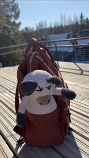 Мягкая игрушка торчит из небольшого рюкзака на деревянном настиле террасы; на заднем плане виден яркий солнечный день и лесной горизонт.