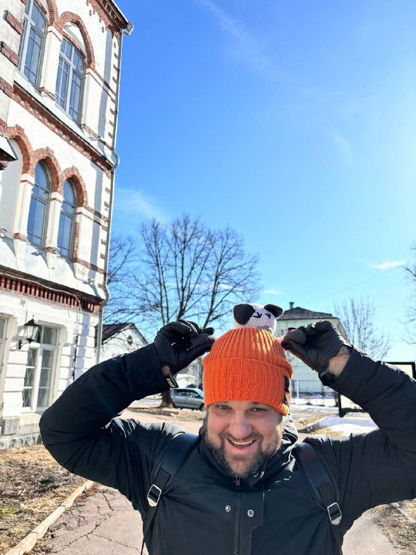 Портретный кадр мужчины в оранжевой шапке и с рюкзаком, он держит игрушку над головой на фоне здания и ясного неба; солнечная прогулка в Сортавале.