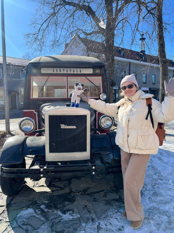Пара на фоне старинного автомобиля у городской набережной; мужчина и женщина позируют рядом с игрушкой, прикреплённой к передней части машины.