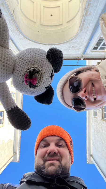 Низко снятый кадр: мужчина и женщина смотрят в камеру, над ними открытое синее небо и элементы архитектуры; связь путешествия и мелочей в кадре.