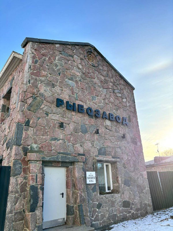 Каменное строение с большой вывеской «РЫБОЗАВОД» на фоне снежного участка; вход небольшого магазина при заводе виден крупным планом.