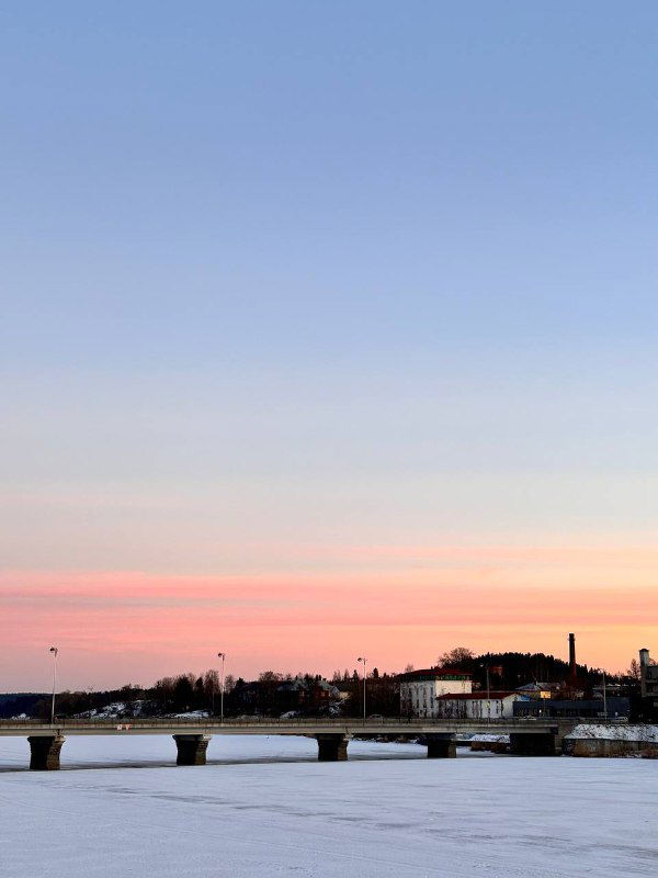 Пейзаж набережной в сумерках: мост через замёрзшую часть залива, низкие здания и мягкие холодные тона заката над горизонтом.