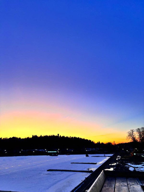 Широкий кадр закатного неба с яркой полосой цвета над линией горизонта и силуэтами деревьев вдоль береговой линии замёрзшего водоёма.