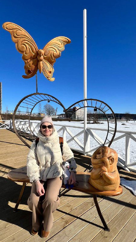 Скульптурная композиция на набережной: большая золотистая бабочка, лавочка и резная фигура, на фоне городской набережной со снегом.