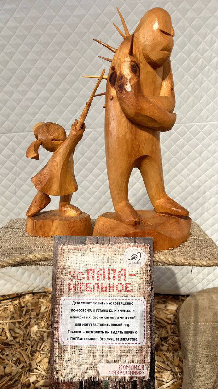 Деревянная скульптура с подписью экспоната: женщина поливает хищное растение, образ заботы и угрозы в одном изображении, табличка рядом.