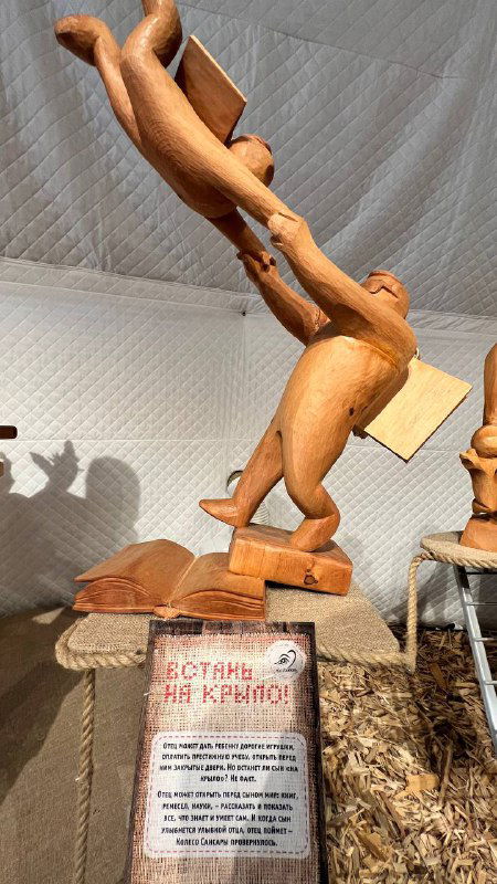 Динамичная деревянная скульптура: фигуры в полёте, подпись экспоната внизу и выставочный зал со мягкими шторками на заднем плане.