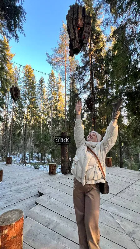 Путешественница подпрыгивает, держит кусок коры или ветку над головой, вокруг настилы, хвойный лес и солнечные лучи.