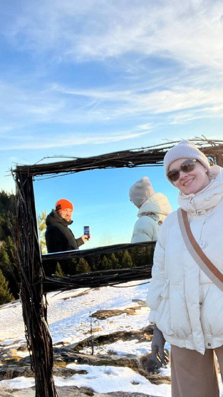 Зеркальная рама на фоне снега и леса, люди стоят у рамки и улыбаются, женщина в светлой куртке у переднего плана.