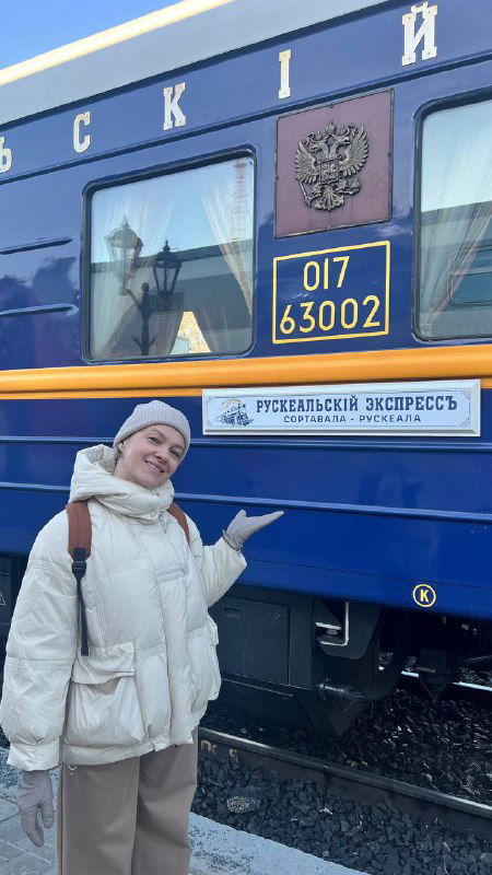 Пассажирка в светлой куртке у синего вагона Рускеальского экспресса, указатель на купе и эмблема на борту на фоне платформы.