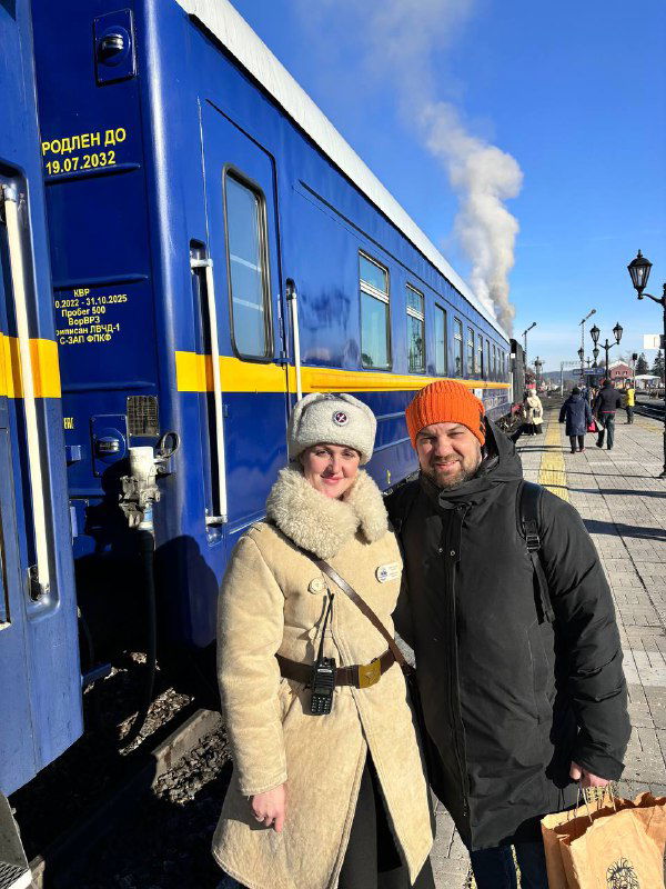 Два человека в тёплой одежде стоят у синего вагона на станции; пар и зимний свет создают праздничную ретро‑атмосферу поездки.