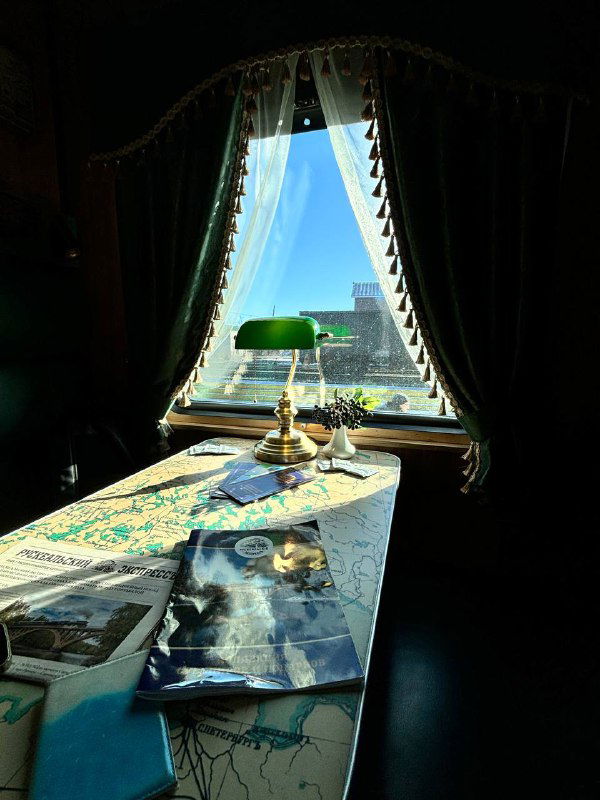 Стол у окна с зелёной лампой, книги и блокнот на столе; вид на морозный пейзаж за стеклом и мягкий интерьер вагона.