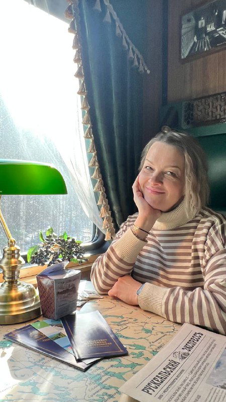 Пассажирка в полосатой кофте улыбается за столиком; дневной свет через окно подчёркивает спокойное настроение поездки.