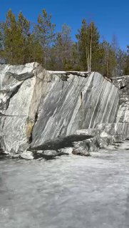 Мраморные стены карьера Рускеала с яркими серыми прожилками, ледяными участками на берегу и солнечным небом над карельской тайгой, весенний день.