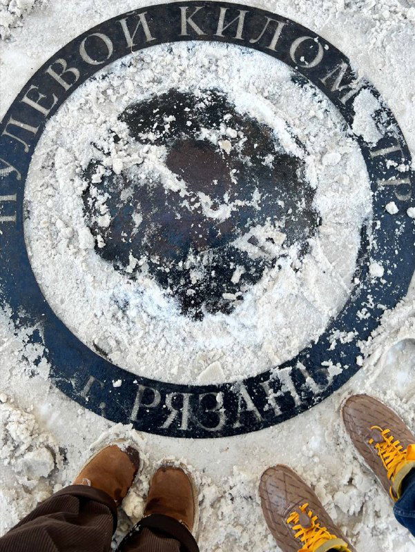 Каменная плита с надписью «нулевой километр Рязани», заснеженная поверхность и ботинки автора в кадре крупным планом.
