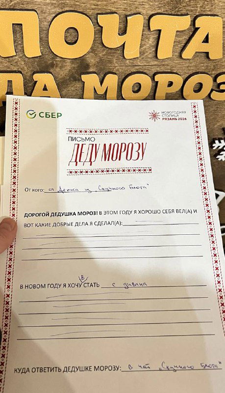 Бланк официального письма Деду Морозу крупным планом на фоне деревянной вывески «Почта Деда Мороза», видно заполненные строки.