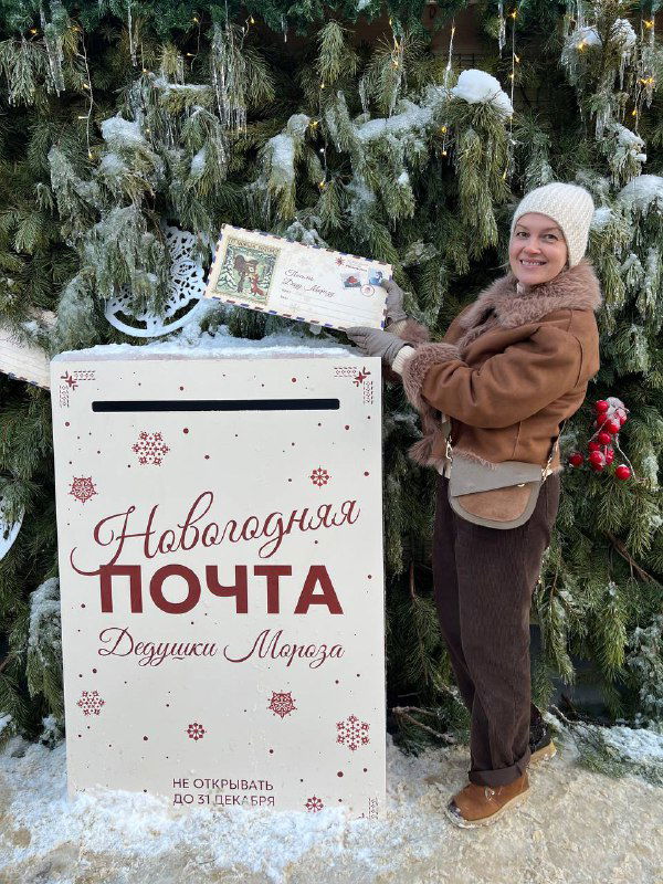 Женщина опускает письмо в праздничный почтовый ящик «Новогодняя почта Дедушки Мороза» на фоне украшенной ели и снега.