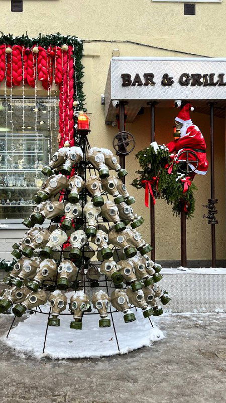 Уличная ёлка, украшенная множеством кружек или чашек, в зимнем городском интерьере рядом с вывеской Bar & Grill и гирляндами.