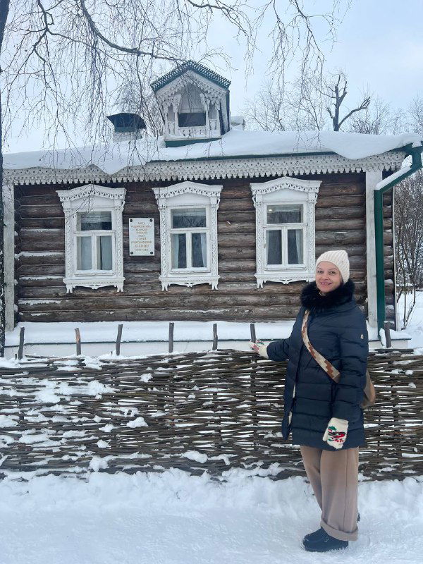 Посетительница на фоне деревянного усадебного дома музея Есенина: женщина в зимней куртке стоит у забора упрятанного в снег домика.