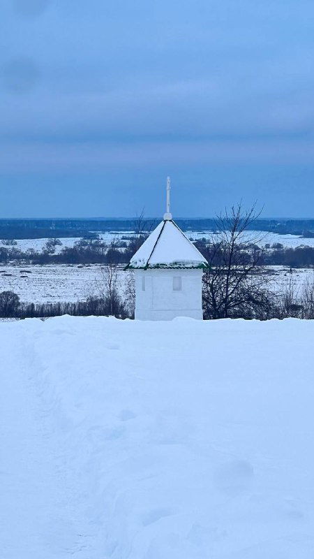 Белая часовня или маленькое купольное здание на фоне снега; тропинка вверху кадра ведёт к строению среди равнины.