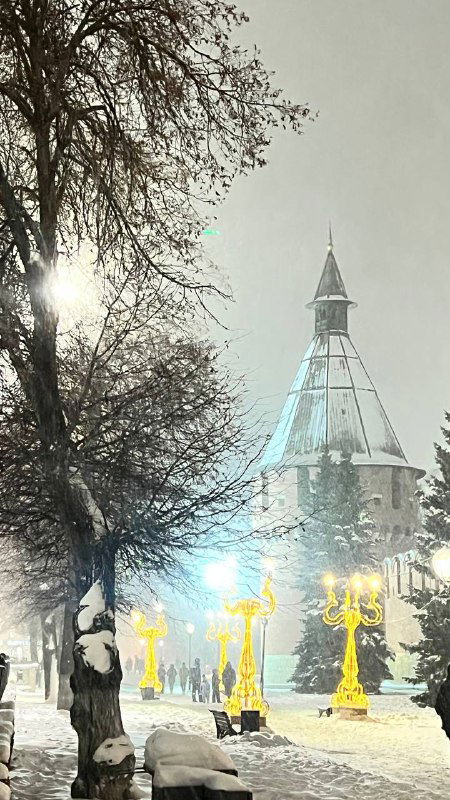 Заснеженная башня кремля или историческое здание Тулы в вечернем свете; деревья, фонари и пухлый снег создают зимнюю картинку города.