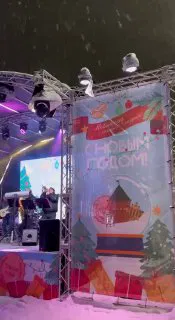 Сцена на площади с праздничным баннером «С Новым годом!» и музыкантами; зрители в тёплой одежде под снегопадом и сценическими огнями.