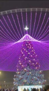 Высокая искусственная ёлка ночью под сиреневым мультимедийным шатром; огни рассеиваются в падающем снегу, создавая праздничную завесу.