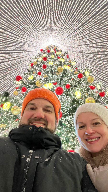 Селфи двух людей на фоне праздничной ёлки и светового купола; улыбающиеся лица в тёплых шапках, снег на одежде и огни позади.