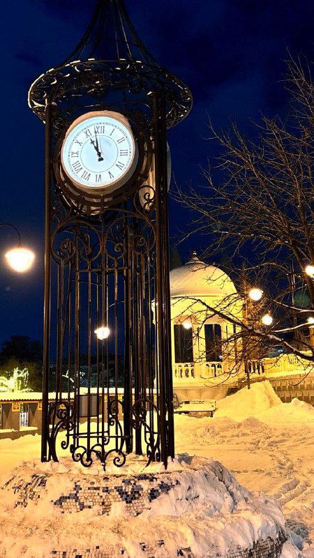 Часы на металлической башенке в ночной Рязани, вокруг снег и огни; урбанистический акцент в праздничном пространстве.