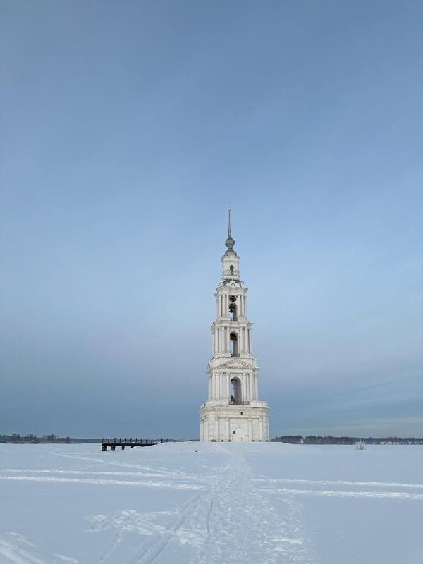 Никольская колокольня Калязина зимой, окружённая льдом и снегом; к ней ведут следы на заснеженной поверхности Волги, бледное небо.
