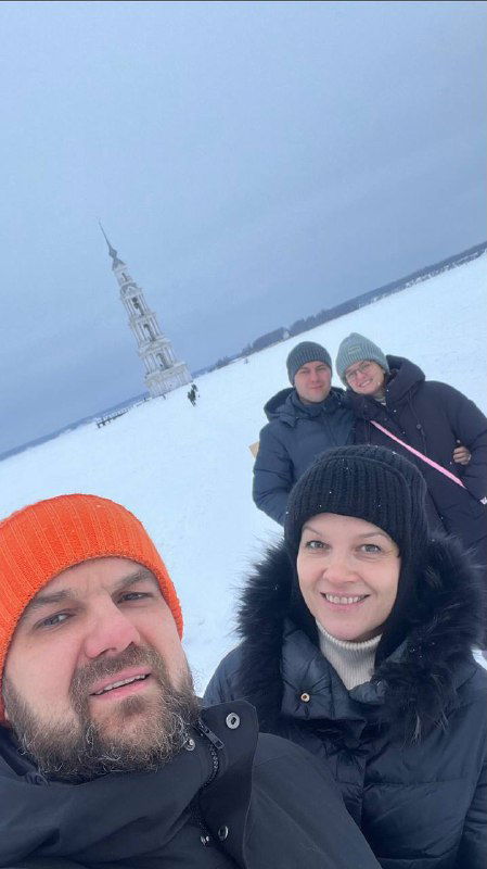 Группа друзей делает селфи на льду Калязина на фоне колокольни; зимой люди тепло одеты, видна прибрежная панорама.