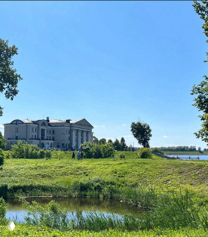 Зелёный берег и историческое здание на фоне летнего неба и реки, пейзаж провинциального города с просторной набережной.