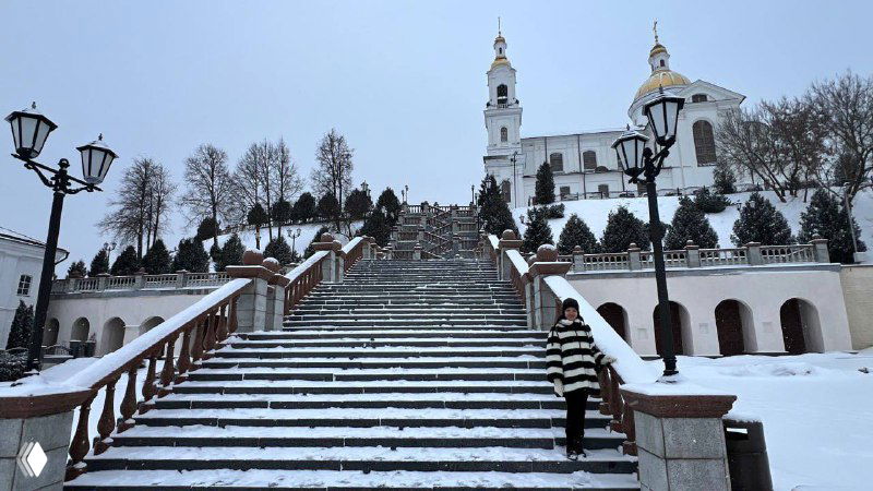 Широкая лестница и панорама Витебска зимой; человек в полосатом пальто поднимается по заснеженным ступеням к храму на холме.