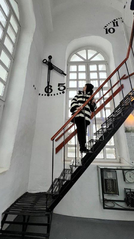 Интерьер ратуши или башни с крупными часами; человек идёт по металлической лестнице внутри белого помещения, видны римские цифры часов.