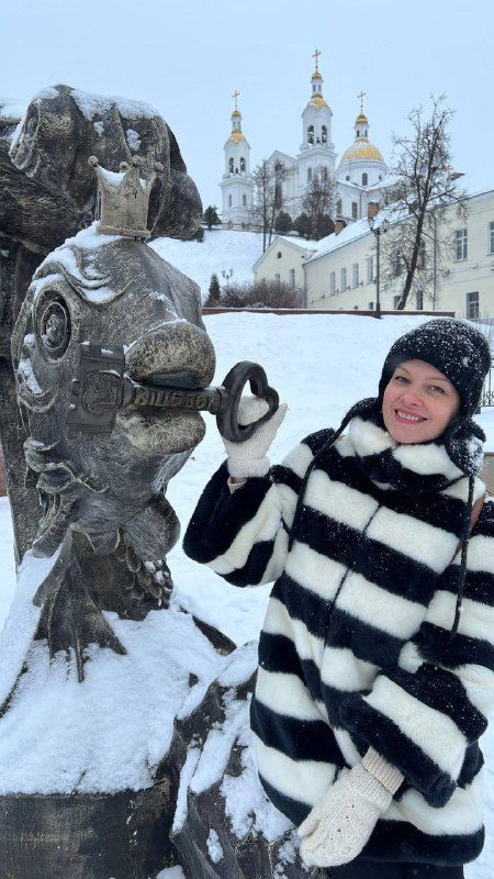 Фигура рыбы-скульптуры и улыбающаяся женщина в полосатом пальто на заснеенной набережной; в фоне видна колокольня и городской пейзаж.