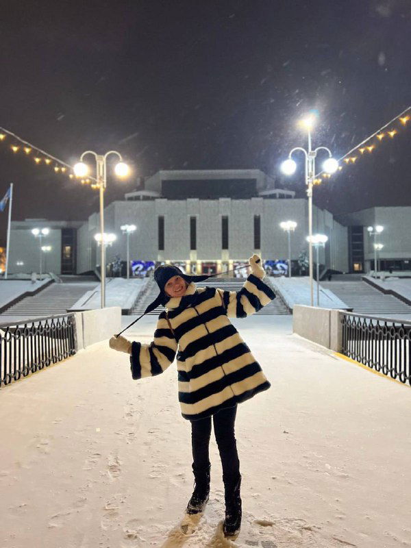 Ночная площадь перед сценой Славянского базара; женщина в пальто позирует на снегу, украшенные улицы и вечерняя иллюминация видны вокруг.
