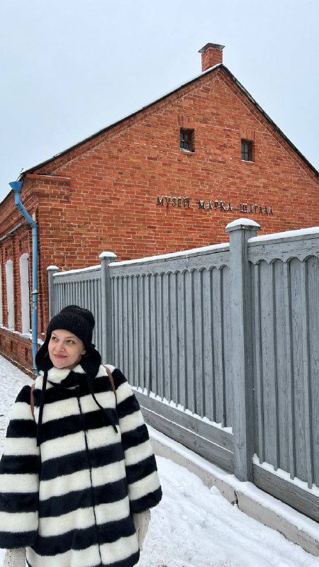 Красный кирпичный дом-музей Марка Шагала с оградой и снегом; на переднем плане женщина в полосатом пальто стоит у забора музея.